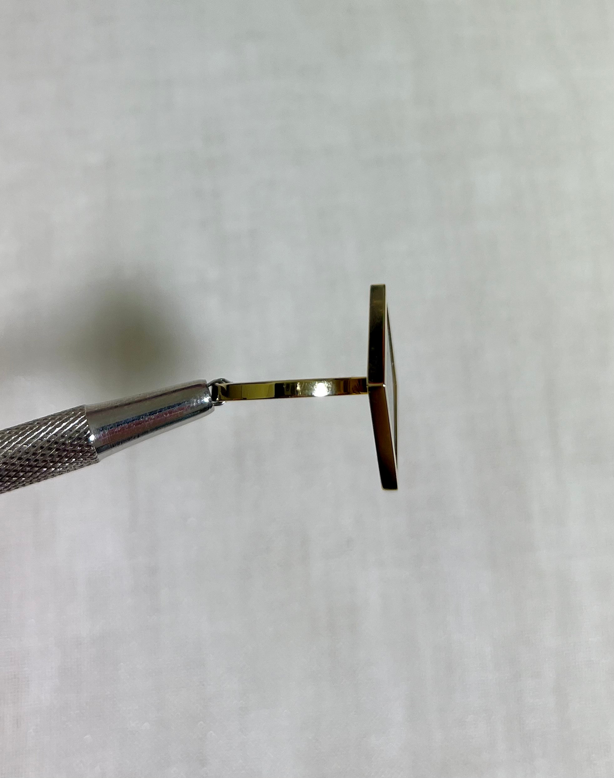 side view of gold square ring held by silver tool on white fabric background 