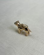 A small polished gold charm shaped like a bulldog, standing on all four legs with a loop at the top, photographed on a light textured fabric background.