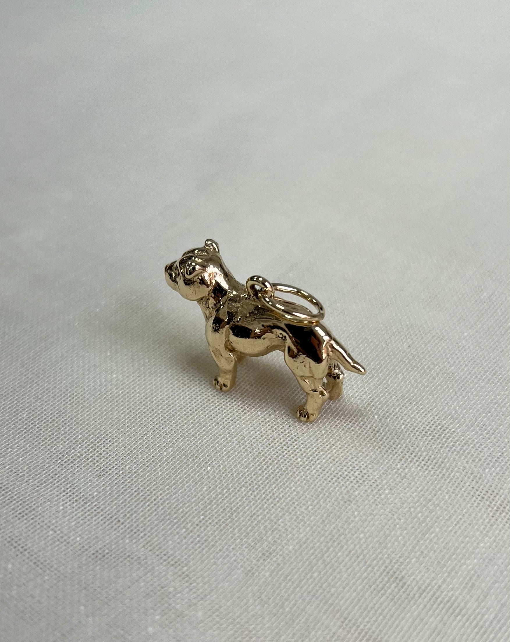 A small polished gold charm shaped like a bulldog, standing on all four legs with a loop at the top, photographed on a light textured fabric background.