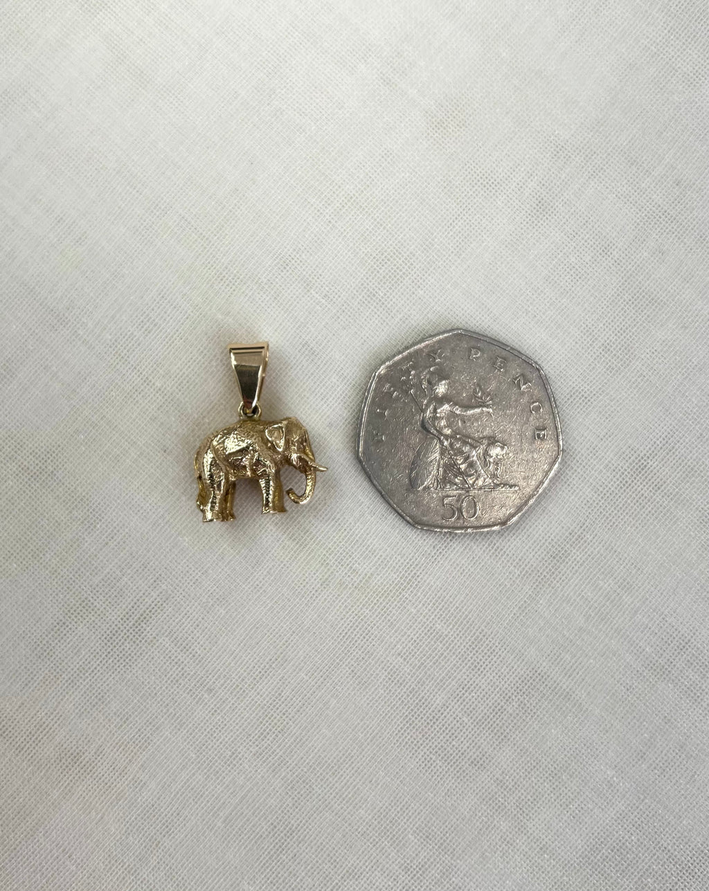 A small gold elephant pendant shown next to a fifty pence coin for scale, placed on a textured off-white fabric background.