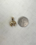 A small gold elephant pendant shown next to a fifty pence coin for scale, placed on a textured off-white fabric background.