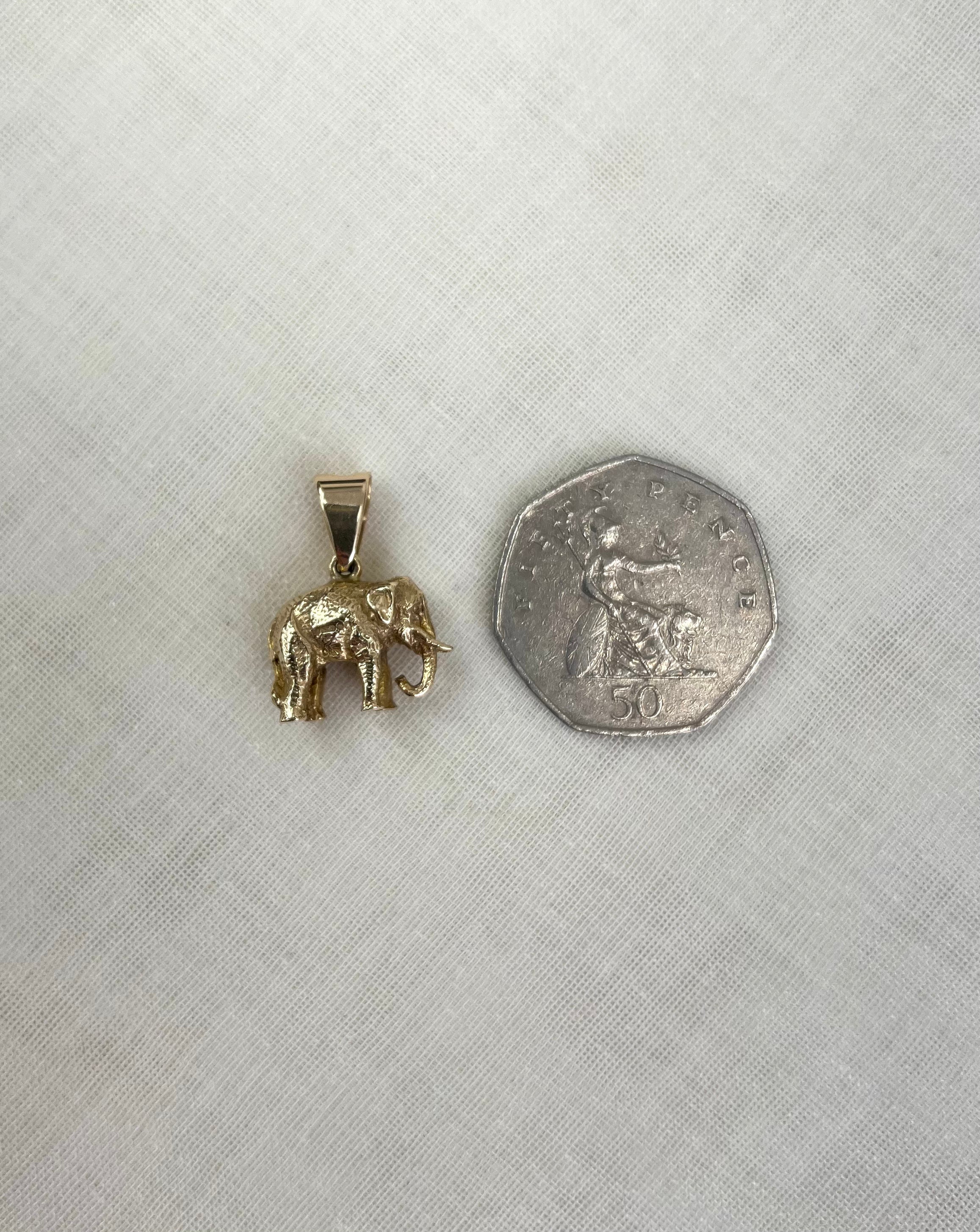 A small gold elephant pendant shown next to a fifty pence coin for scale, placed on a textured off-white fabric background.