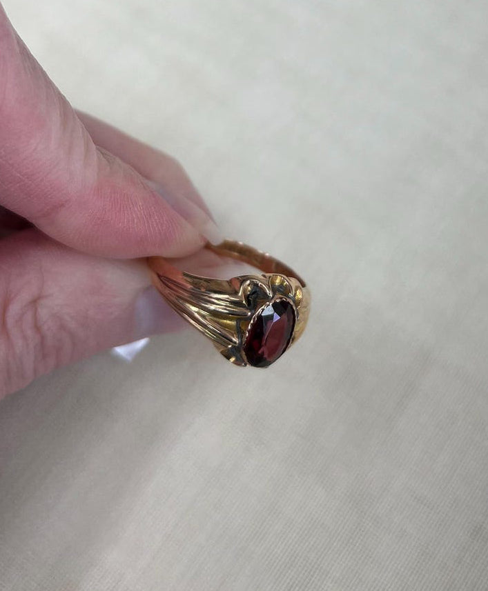 A gold ring with a dark red oval gemstone held between fingers against a light beige fabric background.
