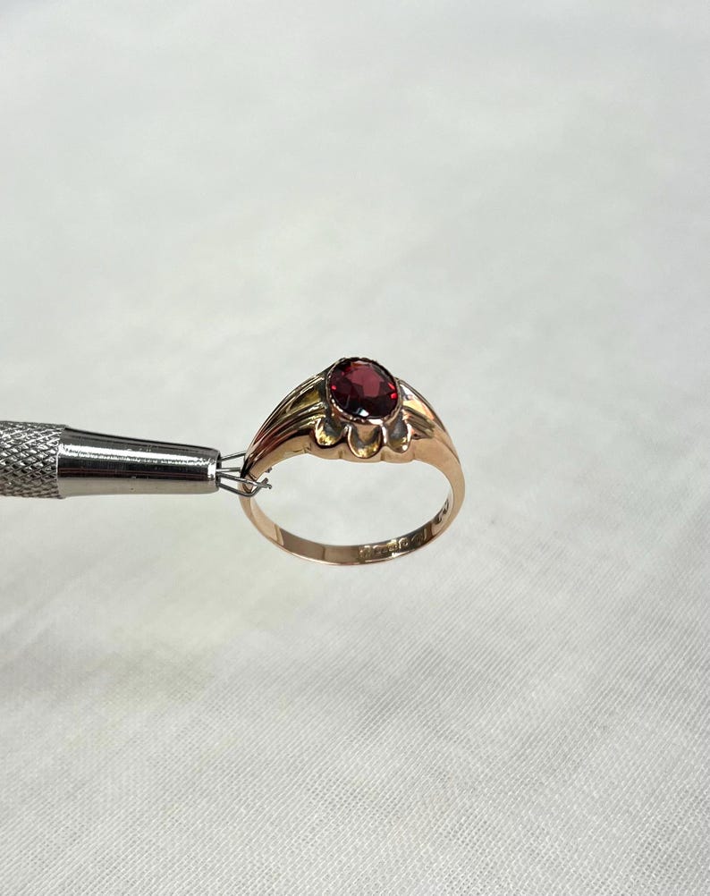A gold ring with an oval red gemstone, held by a silver jeweler's tool against a white textured fabric background.