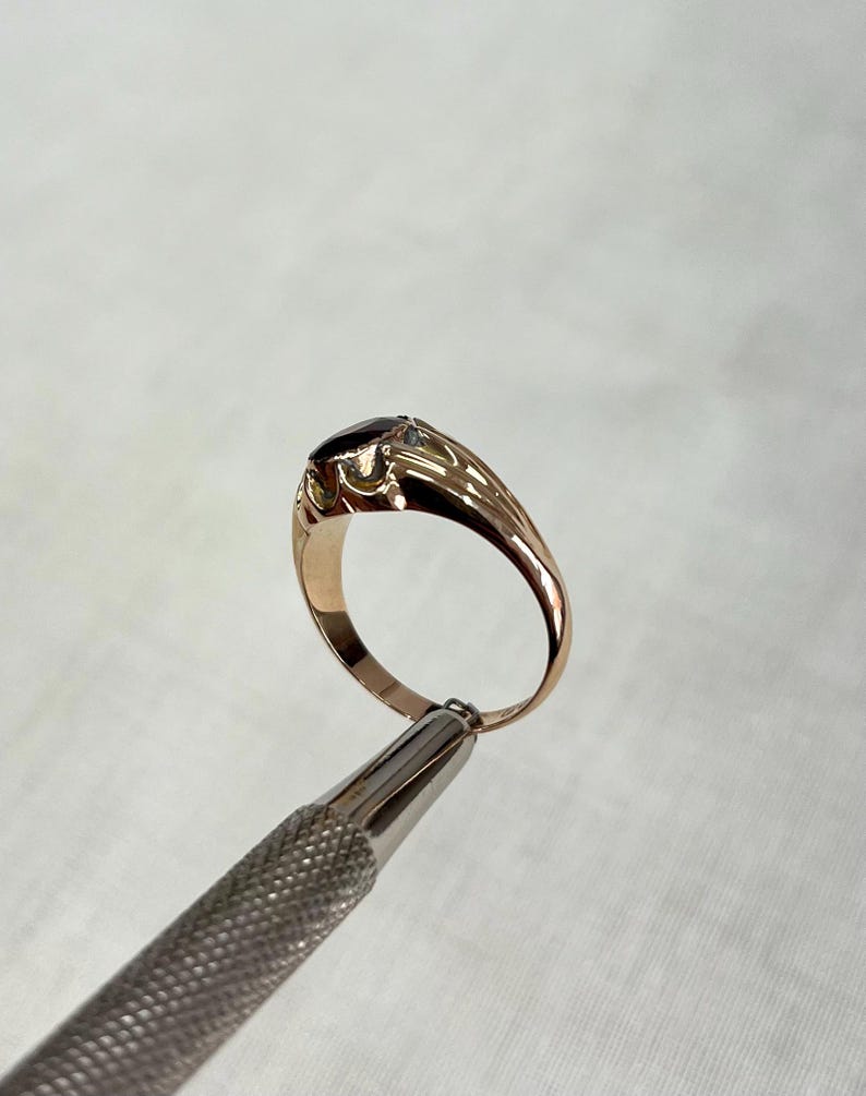 A gold ring with a red oval gemstone, held by a metal tweezer against a light gray textured background.