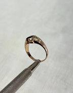 A gold ring with a red oval gemstone, held by a metal tweezer against a light gray textured background.