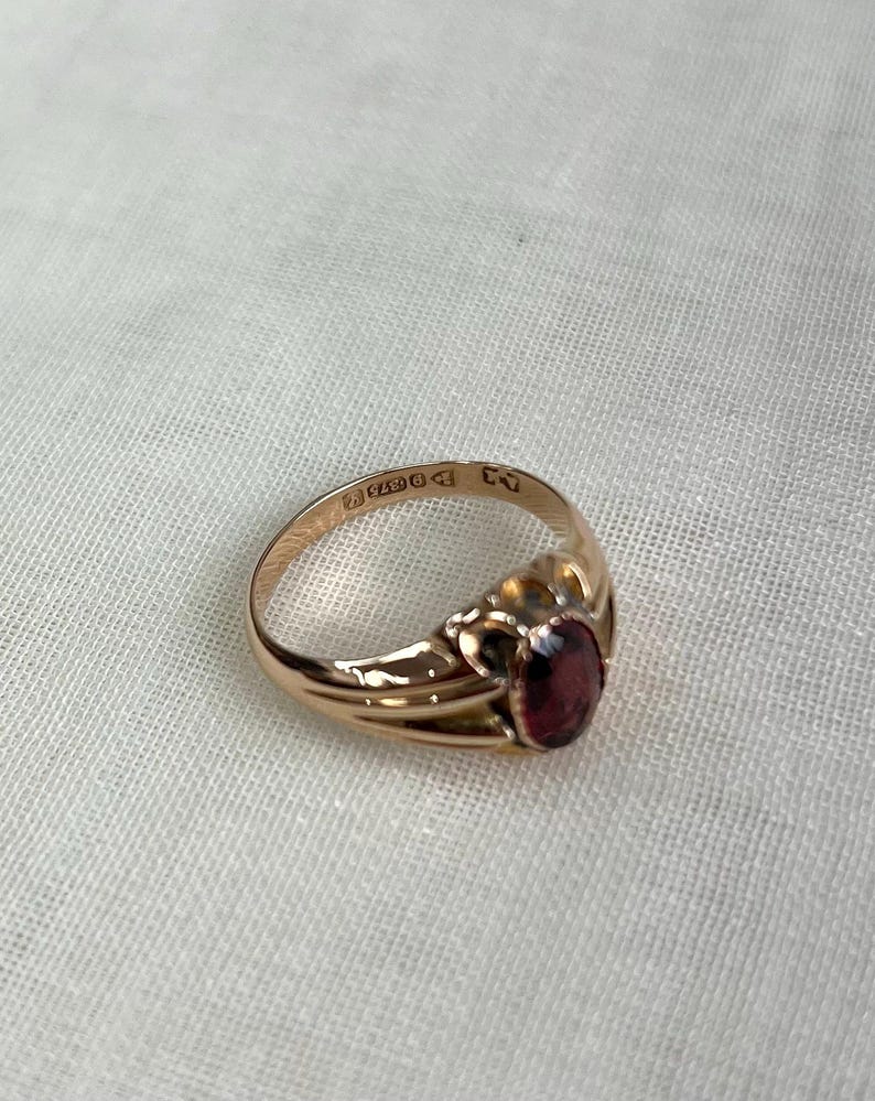gold ring with red gemstone and visible hallmark in focus, resting on textured off white background