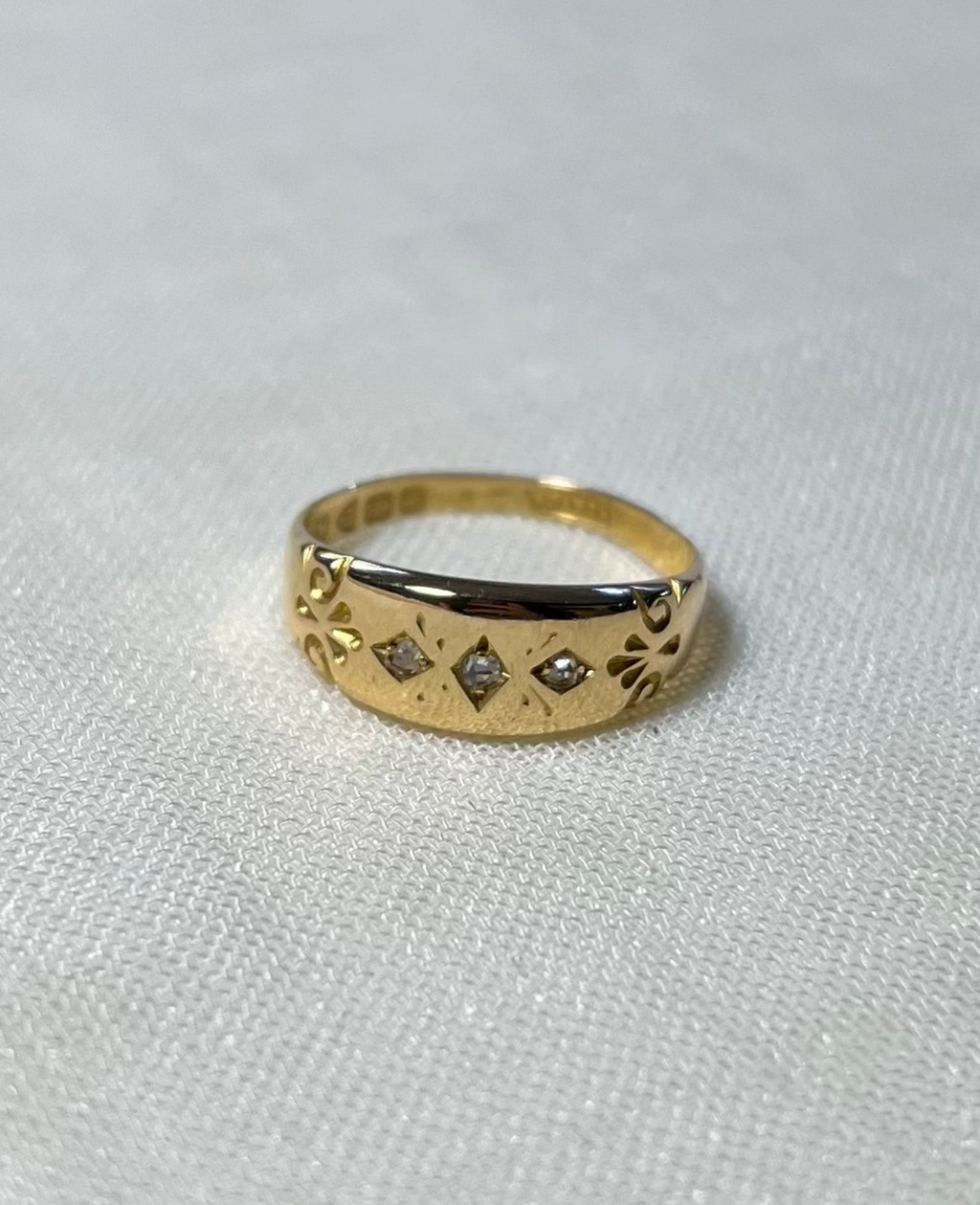 a vintage yellow gold ring with engraved pattern shoulders and 3 diamonds in centre, resting on white fabric background 