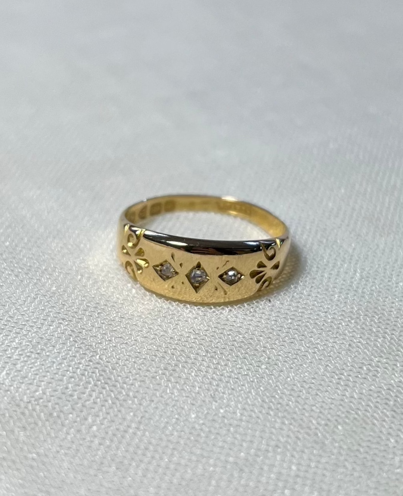 a vintage yellow gold ring with engraved pattern shoulders and 3 diamonds in centre, resting on white fabric background 