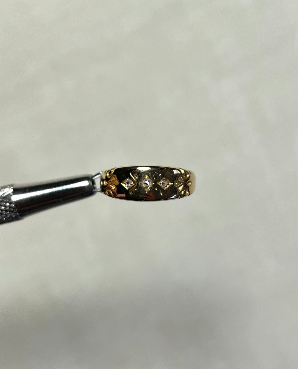A close-up of a small gold ring held by metal tweezers against a soft white background.