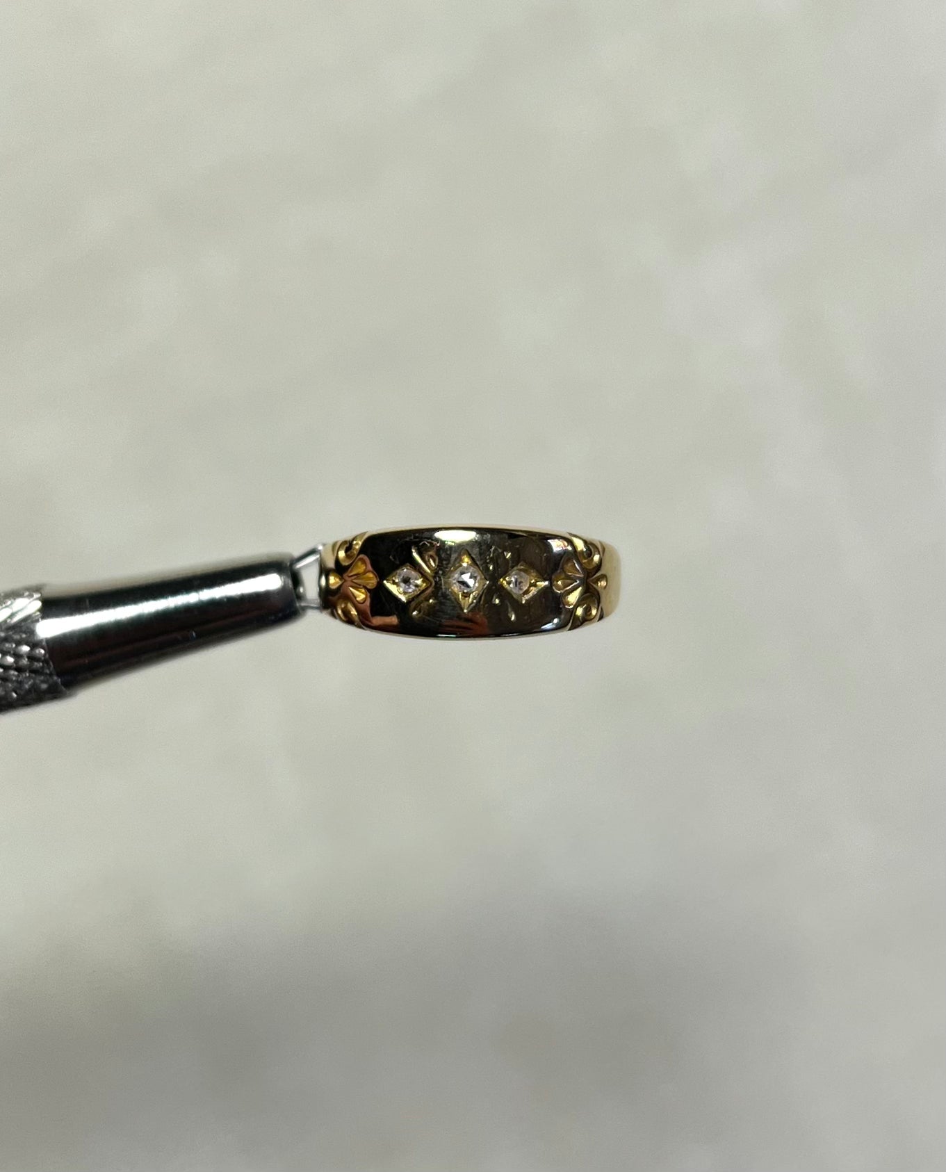 A close-up of a small gold ring held by metal tweezers against a soft white background.