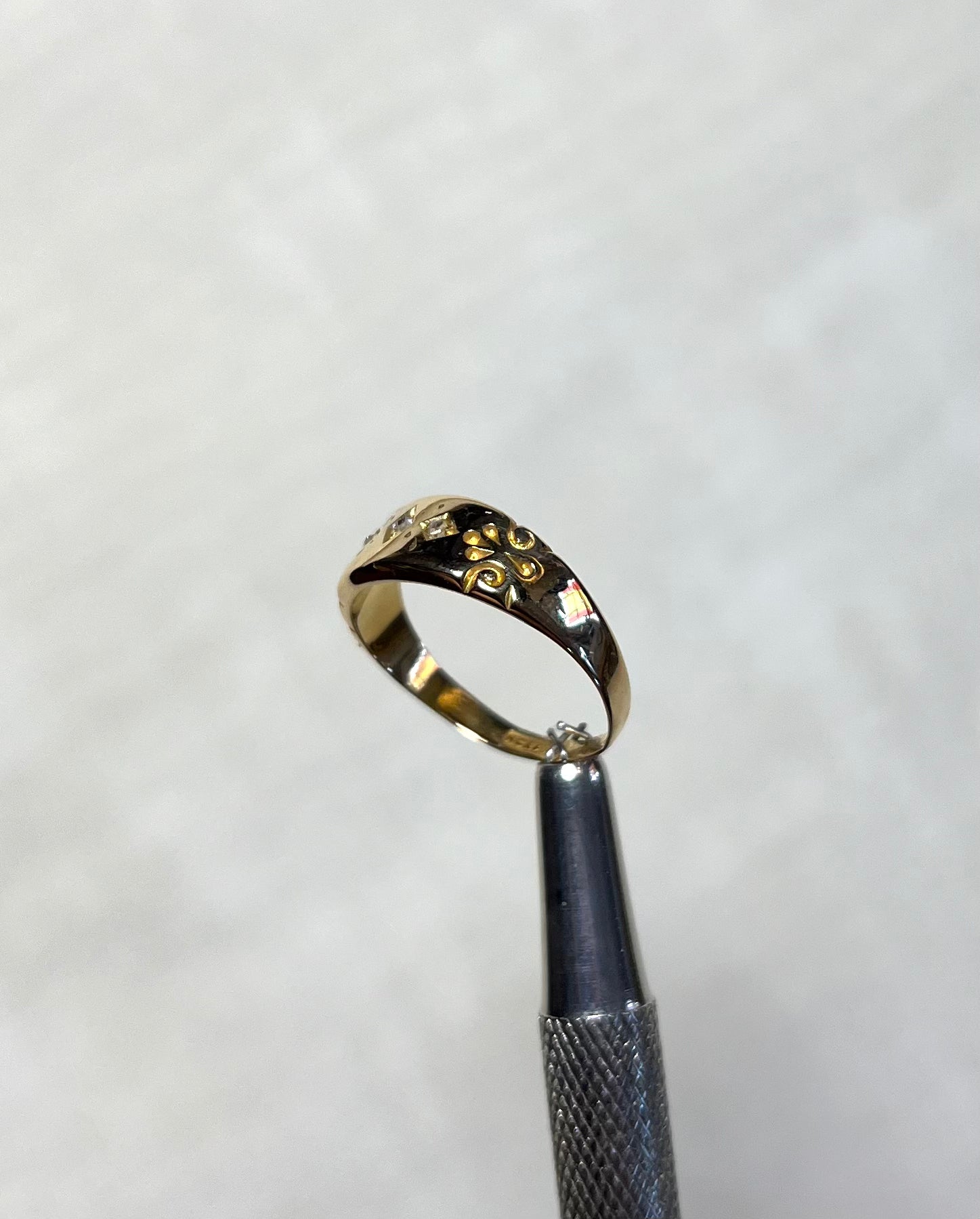 side of gold ring with engraved swirl pattern, held by silver metal tool above soft white background 