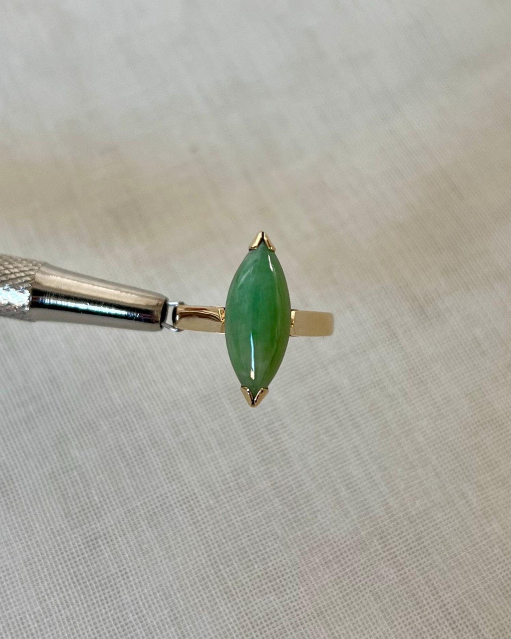 gold ring with marquise shaped green jade stone, held by silver tool above light beige fabric background