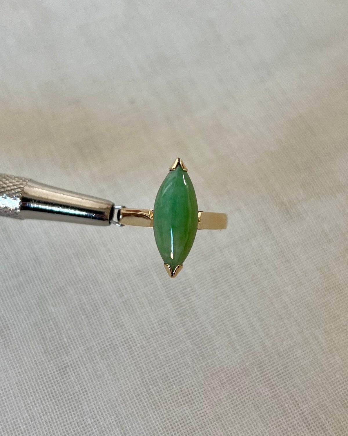 gold ring with marquise shaped green jade stone, held by silver tool above light beige fabric background