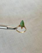 gold ring with green jade stone held by silver tool above light beige fabric background
