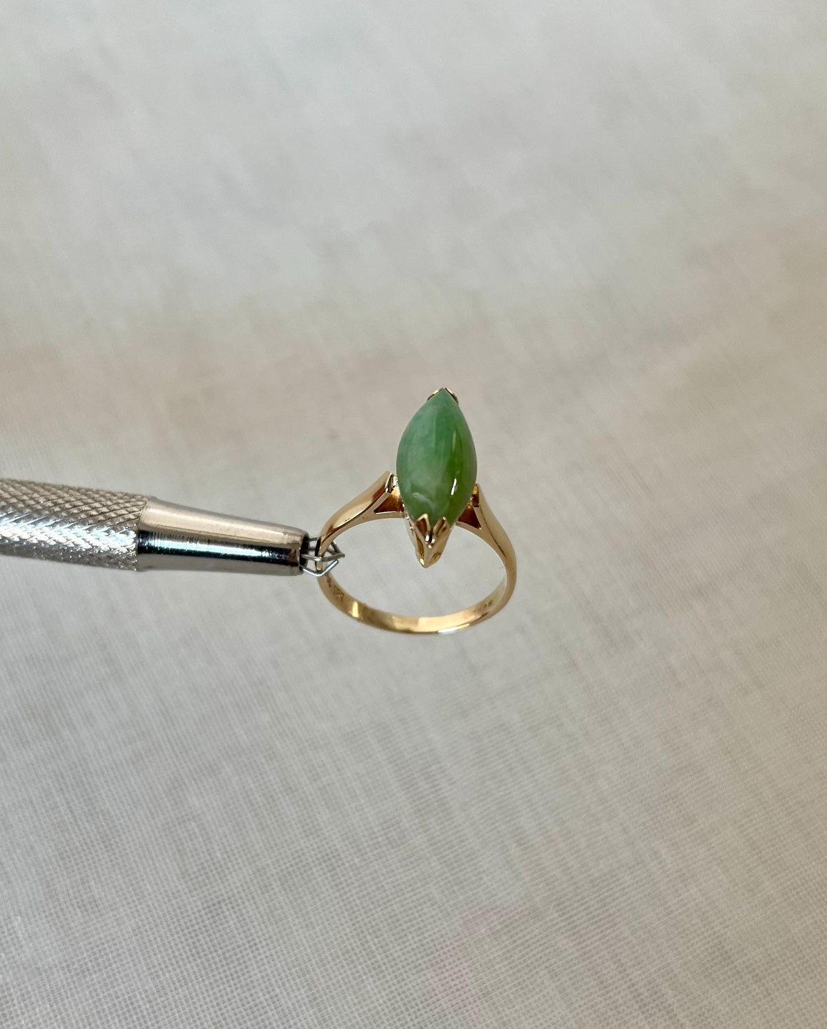 gold ring with green jade stone held by silver tool above light beige fabric background