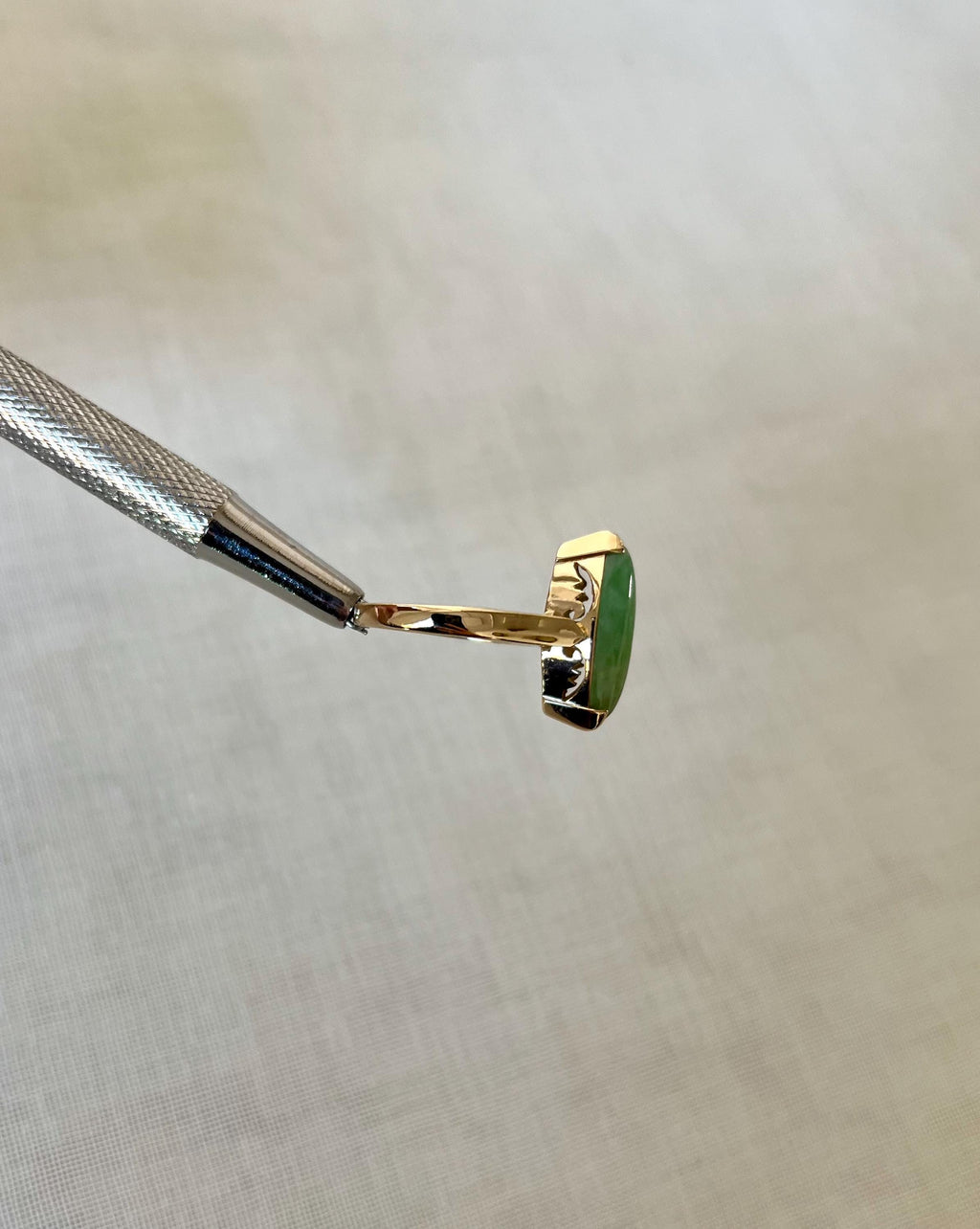 side view of gold ring with green stone, help by silver tool above light beige fabric background 