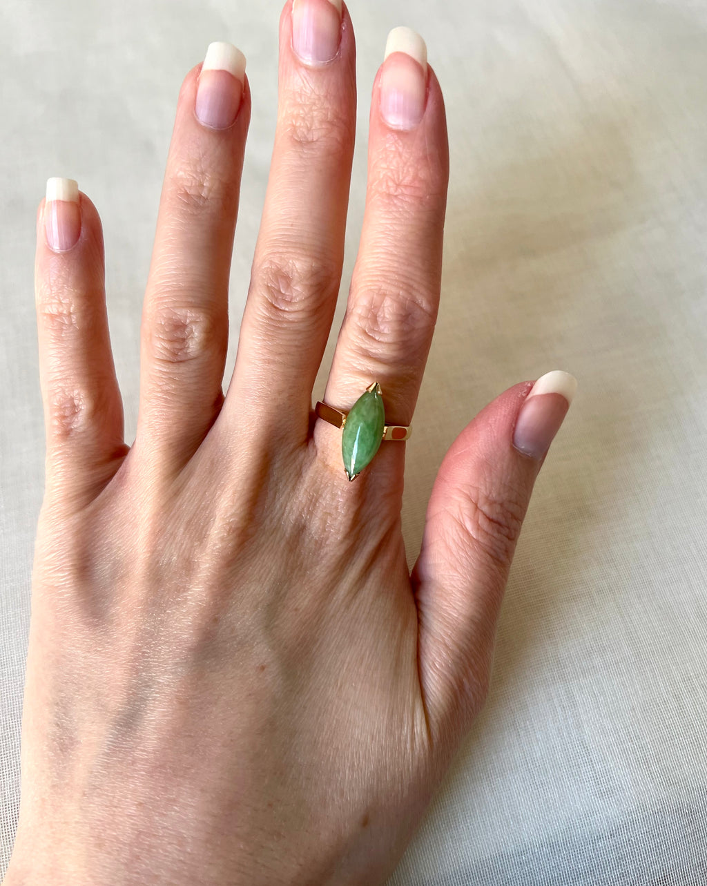 A gold ring with a marquise-cut green gemstone worn on a person's finger, shown against a light beige fabric background.