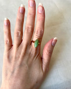 A gold ring with a marquise-cut green gemstone worn on a person's finger, shown against a light beige fabric background.