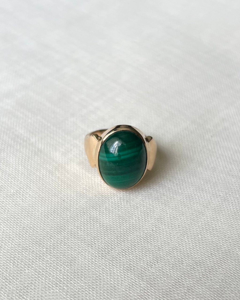 A gold ring with an oval green malachite stone, resting on a textured white fabric background.