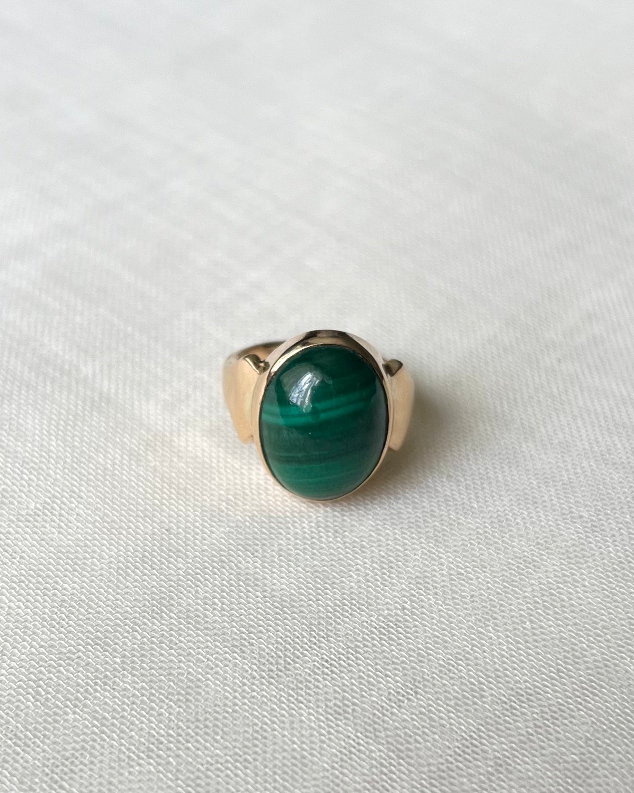 A gold ring with an oval green malachite stone, resting on a textured white fabric background.