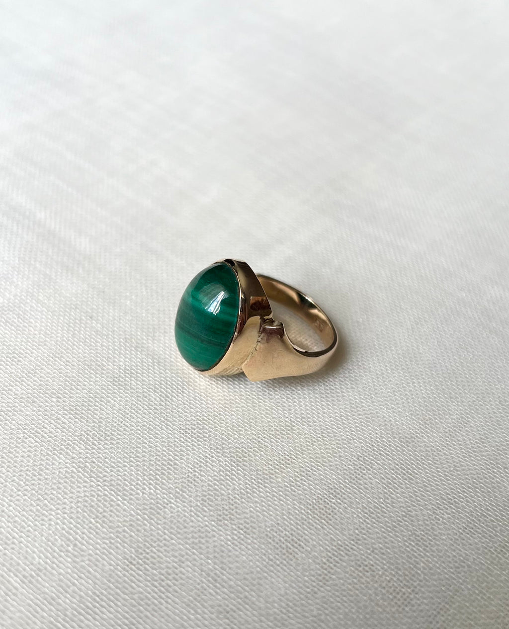 A gold ring featuring an oval-shaped green malachite stone, resting on a textured white fabric background.