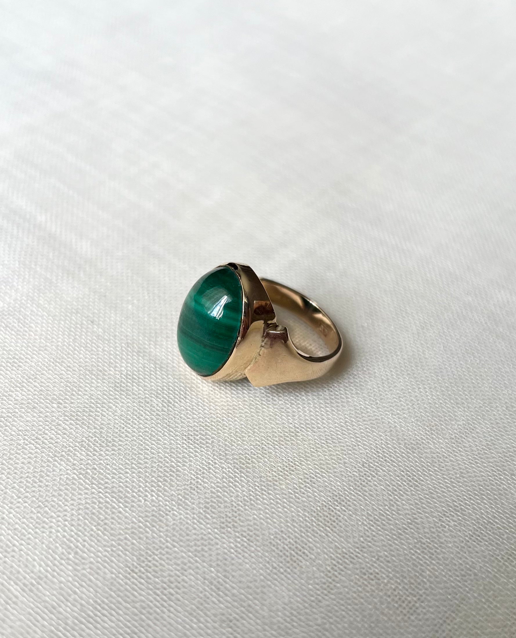 A gold ring featuring an oval-shaped green malachite stone, resting on a textured white fabric background.
