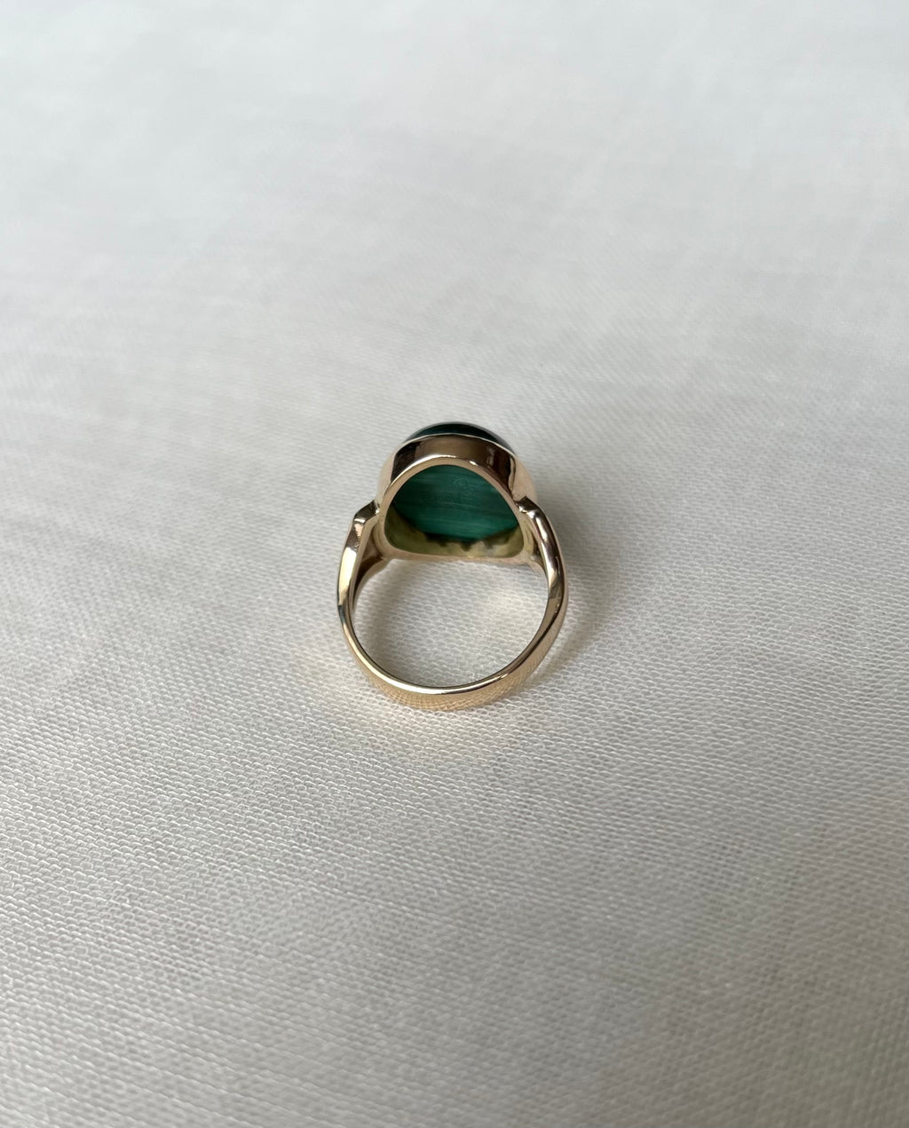 a gold ring with a oval-shaped green stone, photographed from behind on a textured off-white fabric background.