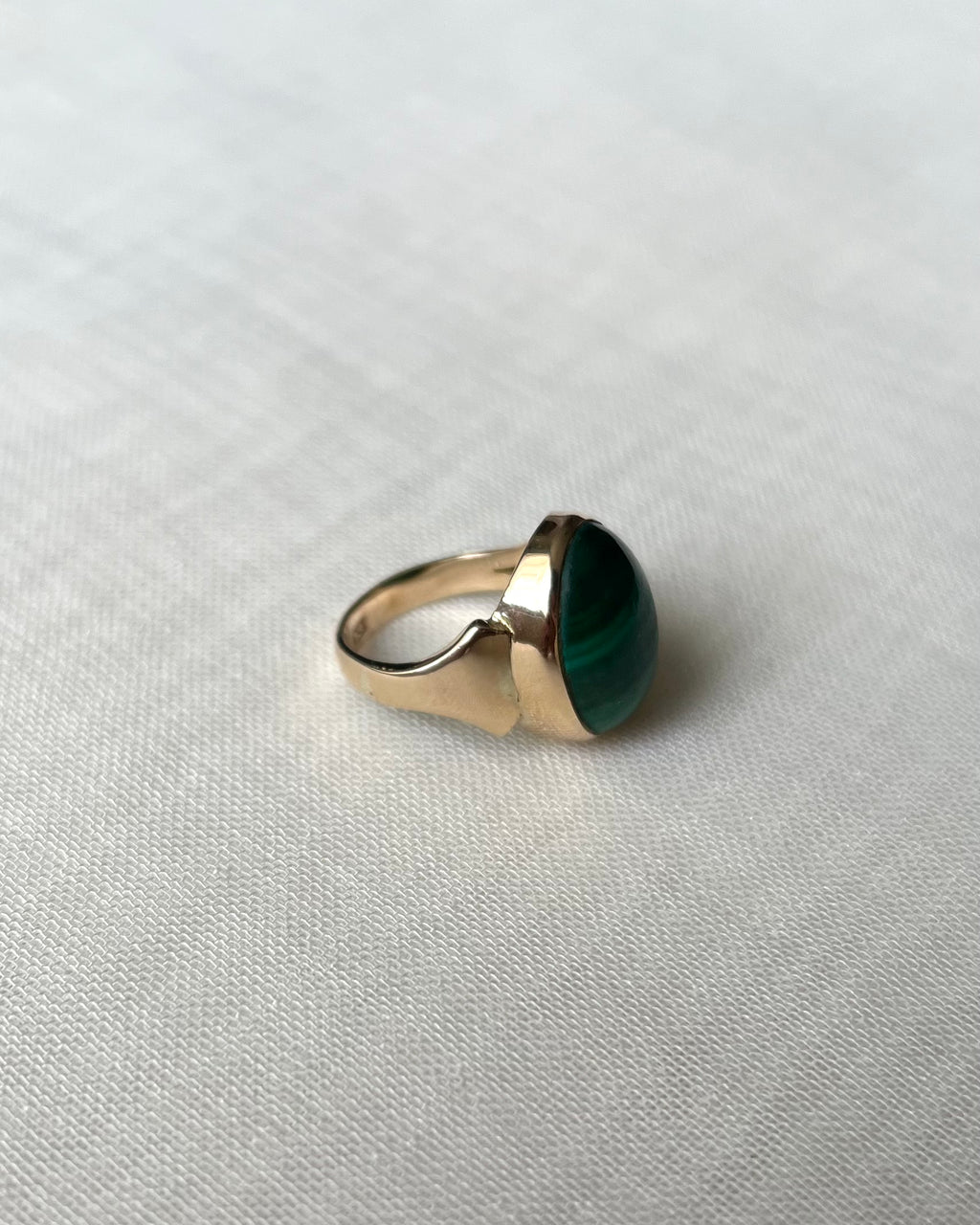 A gold signet-style ring with a large oval green malachite gemstone, resting on a textured off-white fabric background.