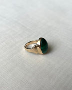A gold signet-style ring with a large oval green malachite gemstone, resting on a textured off-white fabric background.