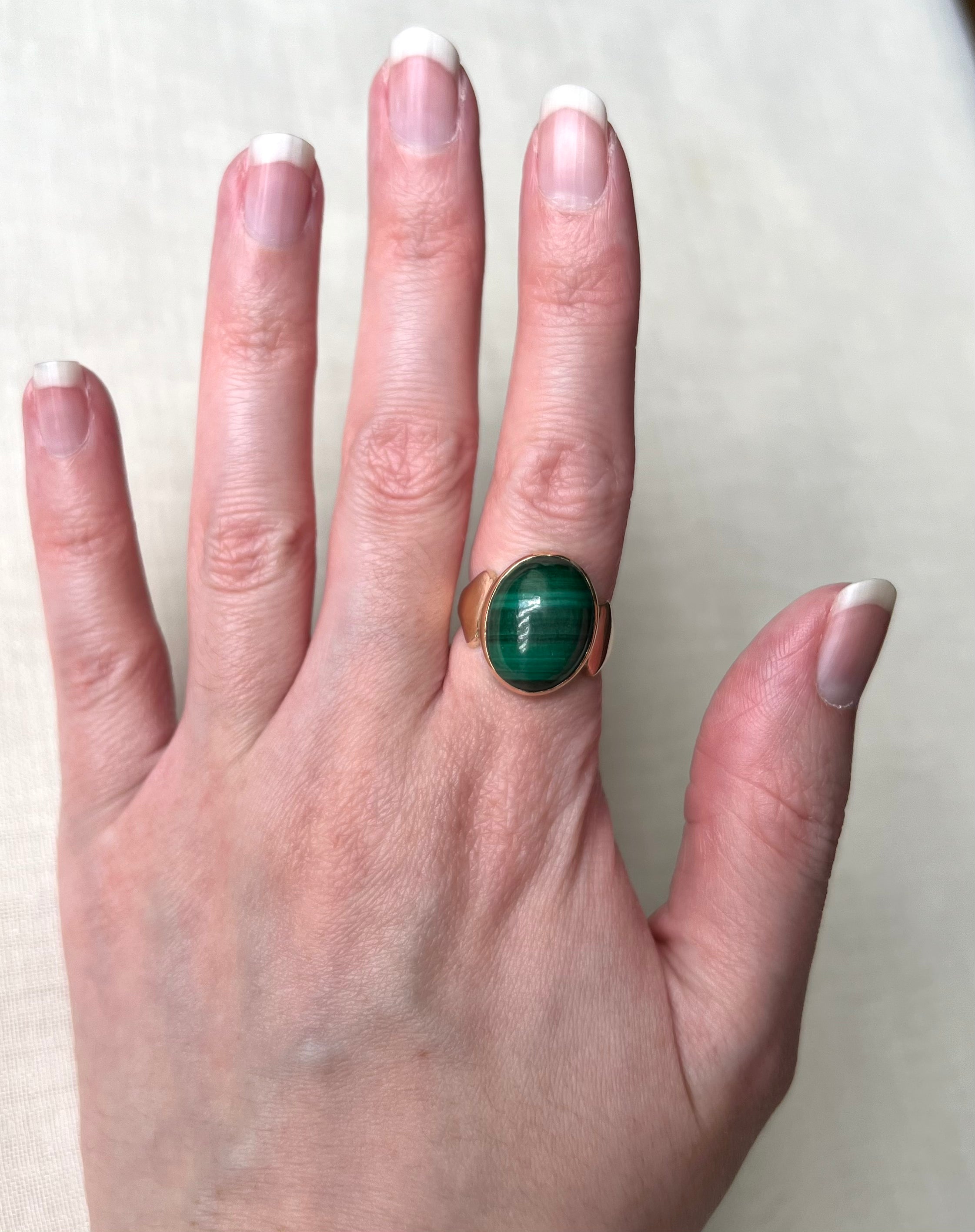 A close-up of a hand wearing a gold ring with a green malachite cabochon stone, set against a neutral light beige background.