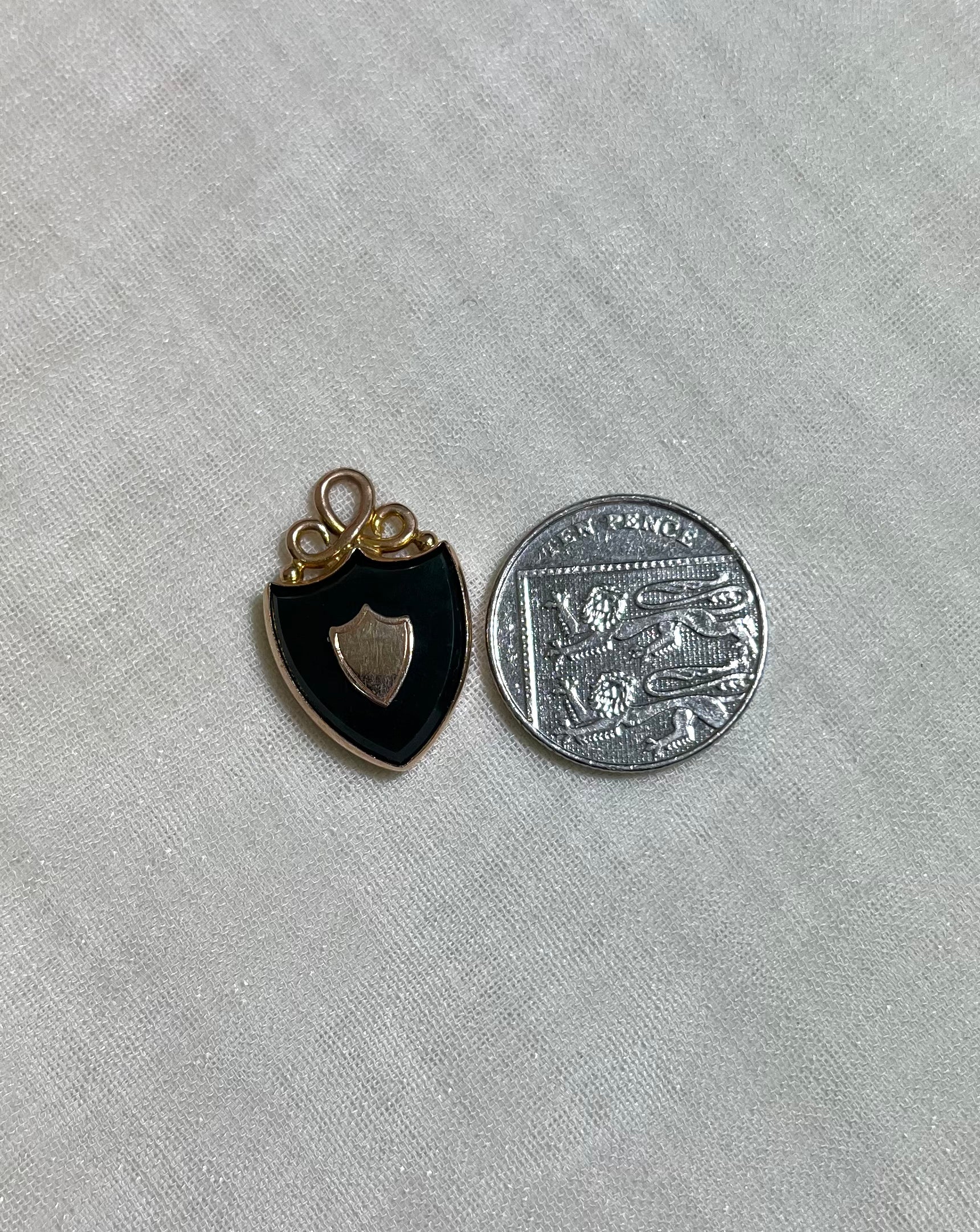 gold shield pendant next to ten pence coin for size reference, on white fabric background