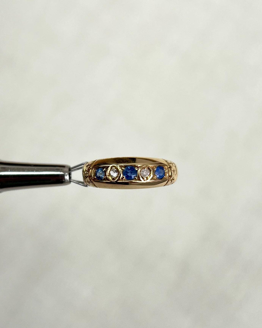 gold ring set with blue sapphires and diamonds, held by silver metal tool above soft white background