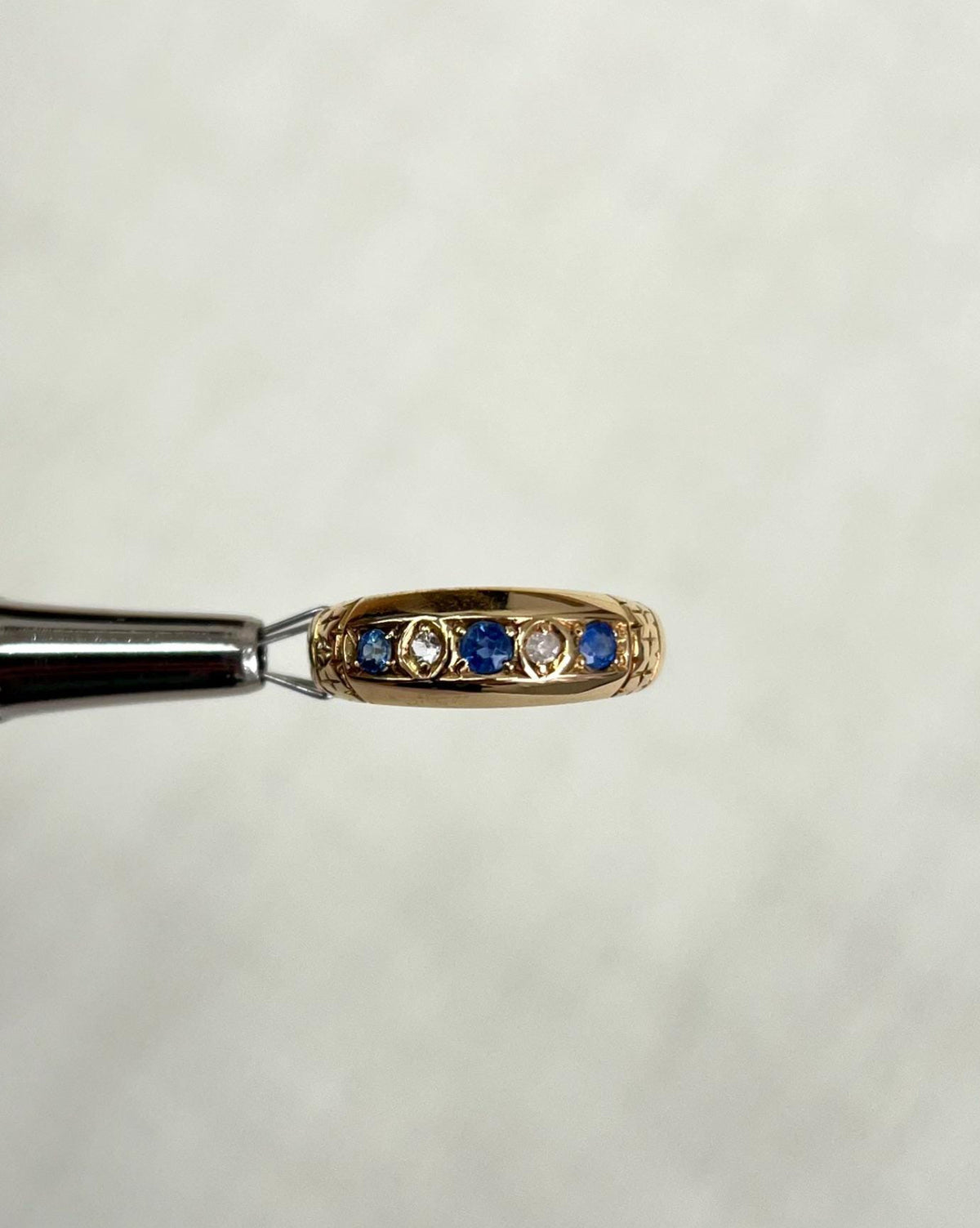 gold ring set with blue sapphires and diamonds, held by silver metal tool above soft white background