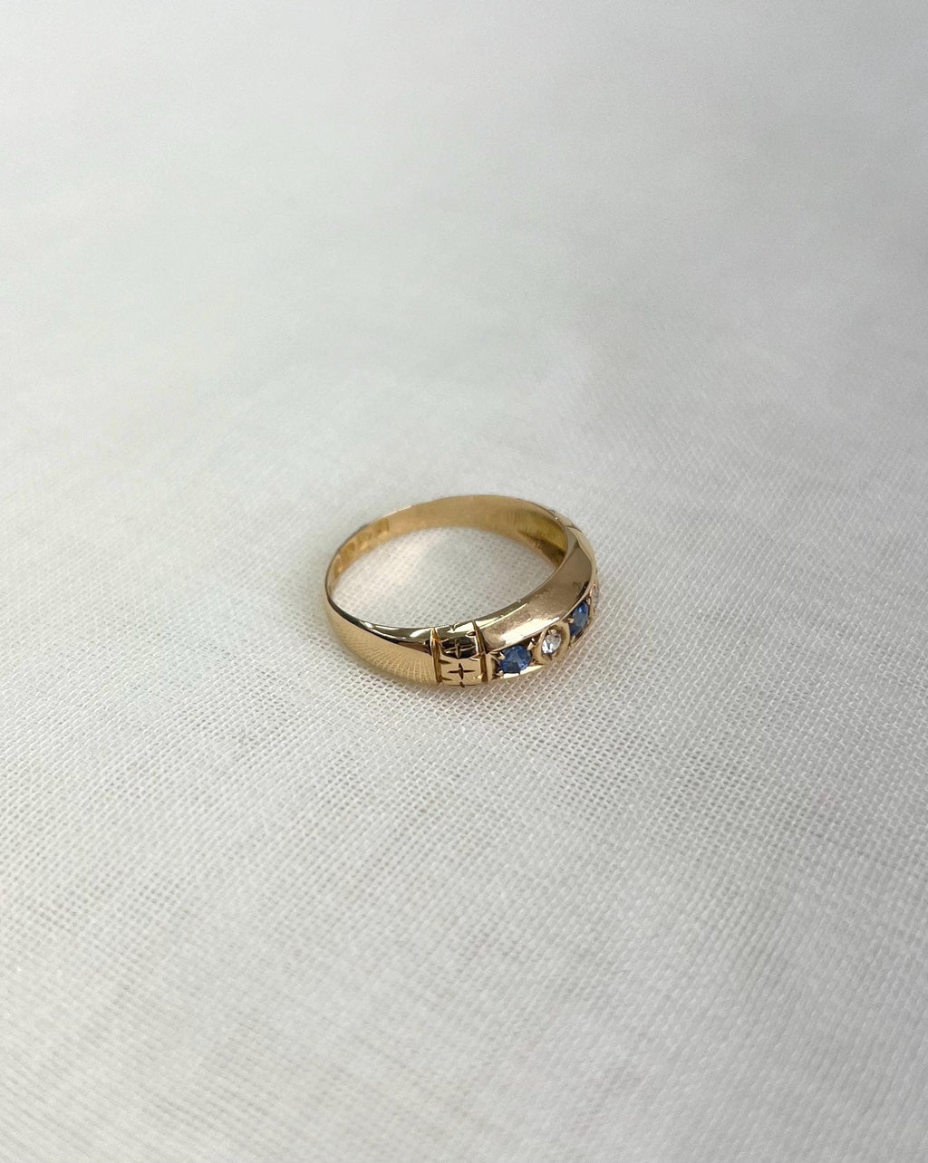 gold band with blue and clear gemstones and shoulder engraving, resting on white fabric background
