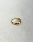 gold band with blue and clear gemstones and shoulder engraving, resting on white fabric background
