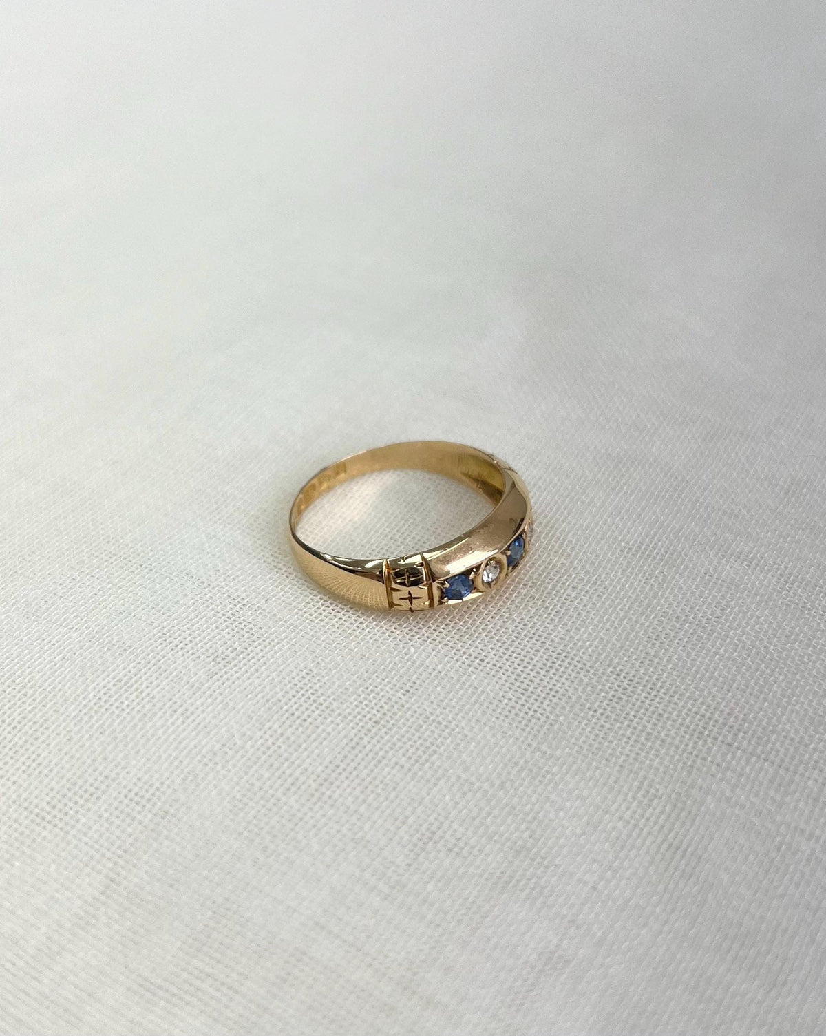 gold band with blue and clear gemstones and shoulder engraving, resting on white fabric background