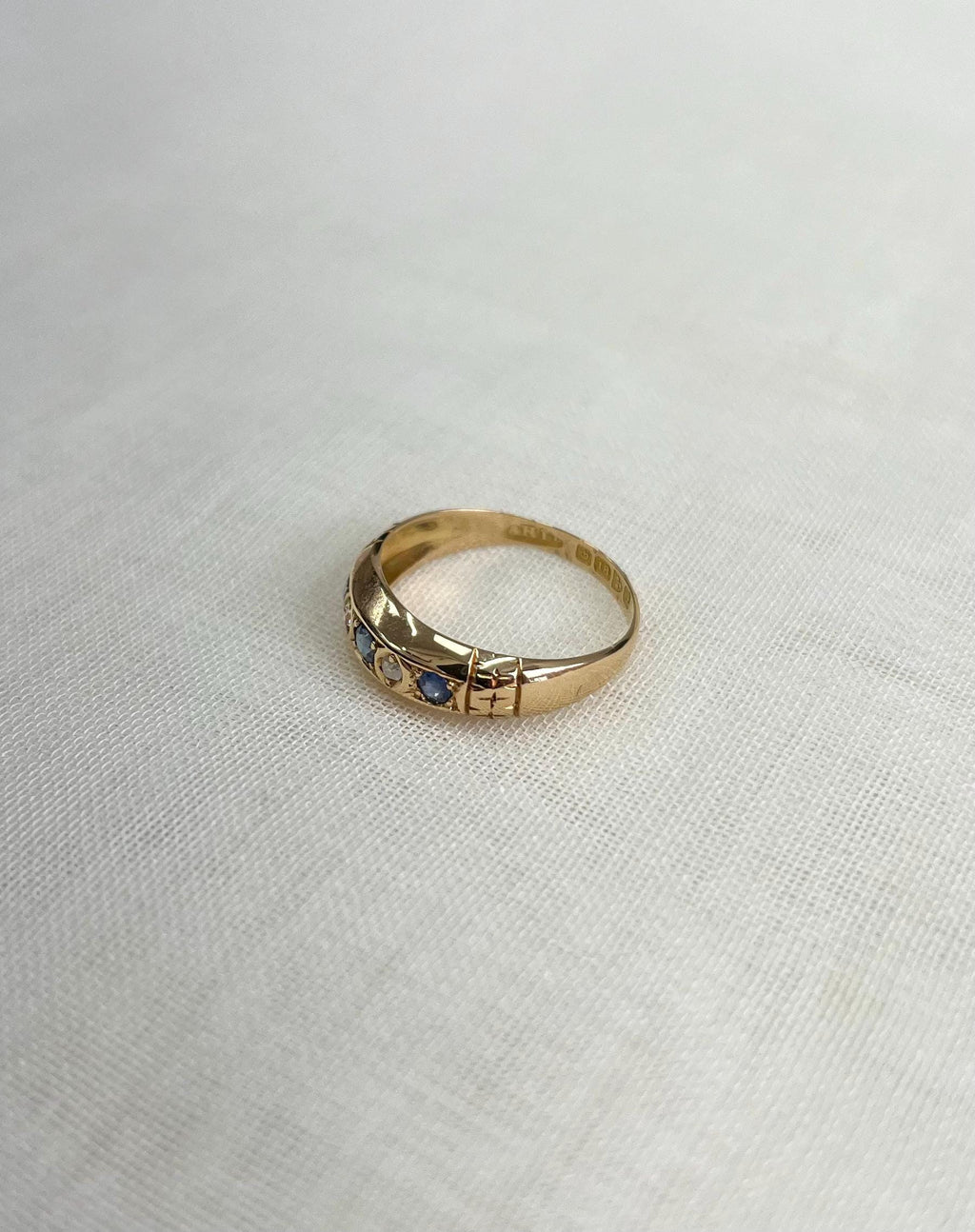 gold ring with blue and clear gemstone and shoulder engraving, resting on white fabric background