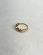 gold ring with blue and clear gemstone and shoulder engraving, resting on white fabric background