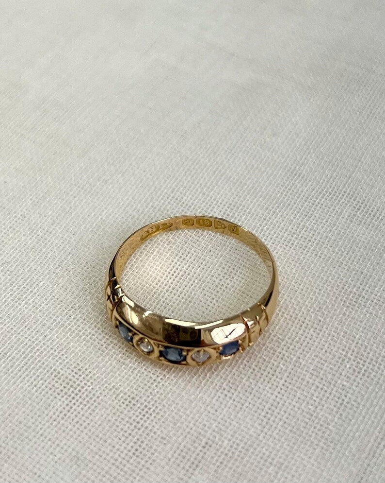 gold rings with blue and clear gemstones with focus on visible hallmark (18, Chester symbol) resting on white fabric background 