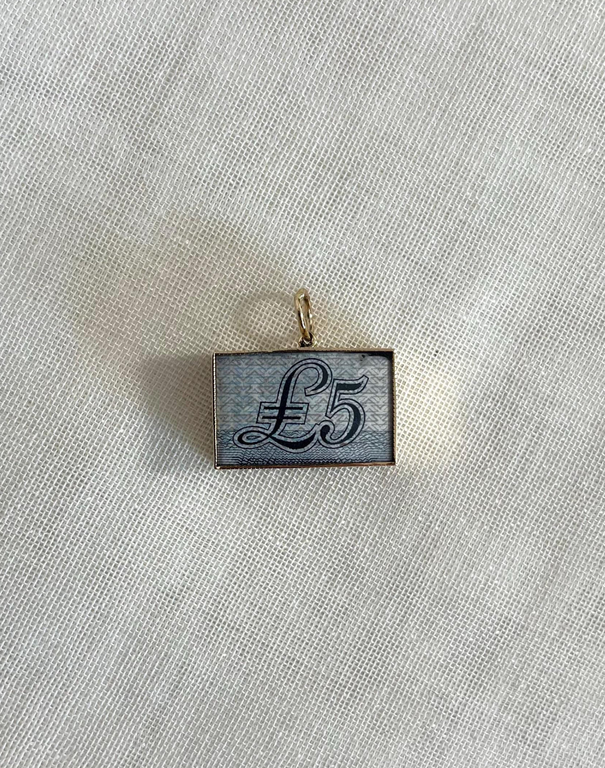 rectangle gold charm with five pound note inside, resting on off white fabric background 