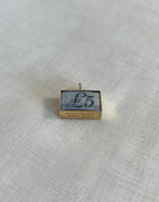 gold rectangle charm with five pound note inside and 'in emergency break glass' inscription on the bottom, resting on off white fabric background