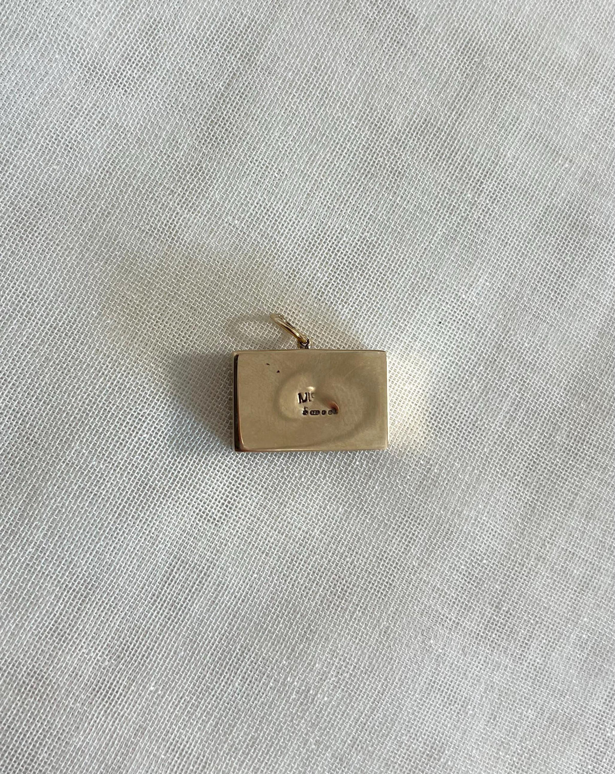 back of rectangle gold charm with hallmark visible, resting on off white  fabric background 