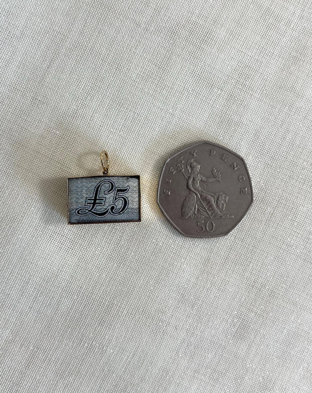 rectangle gold charm with five pound note inside next to 50 pence coin for scale, resting on off white background 