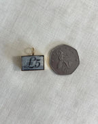 rectangle gold charm with five pound note inside next to 50 pence coin for scale, resting on off white background 