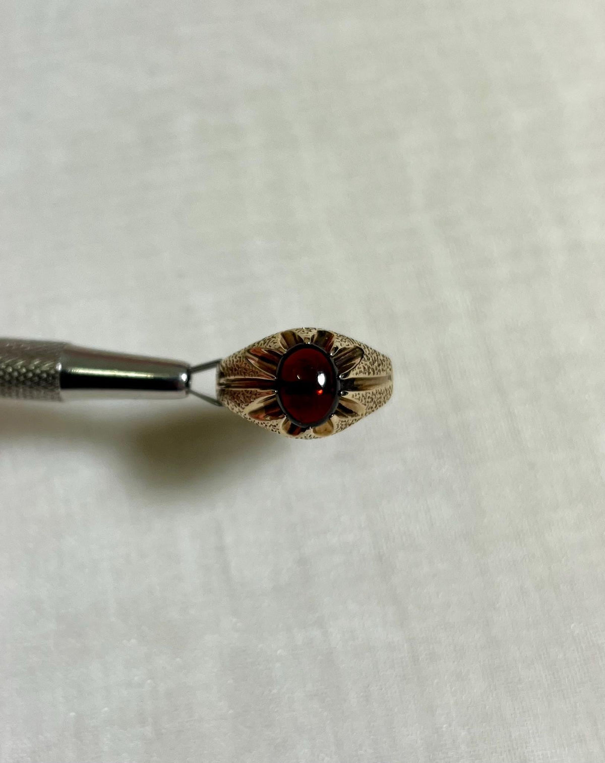 gold ring with textured sides and red stone centre, help by silver tool above cream coloured background 