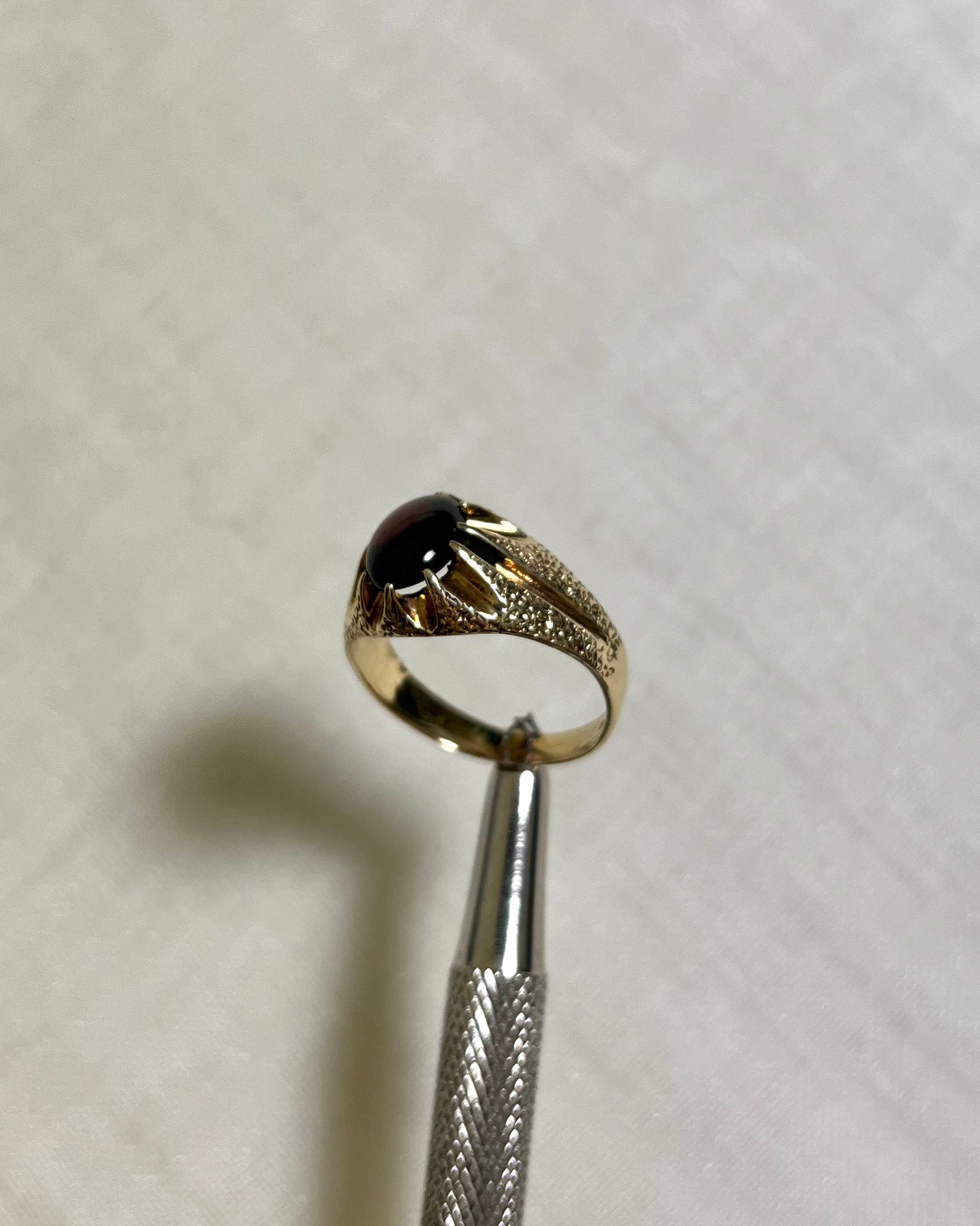 side view go gold ring with red centre stone and textured shoulders, help by silver tool above ight cream background 