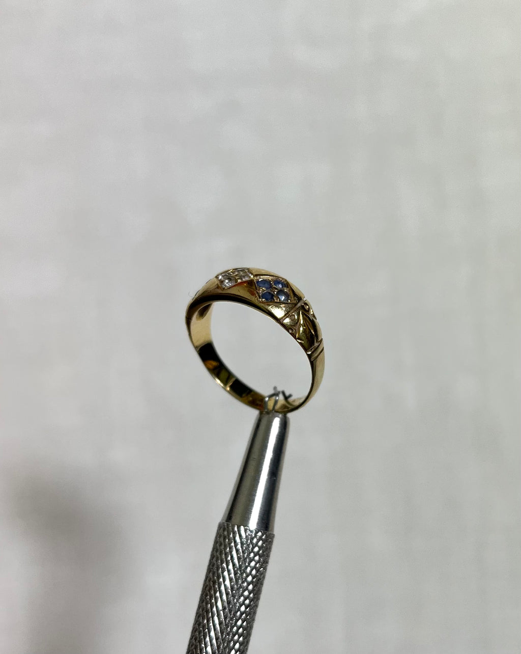 A gold ring with small blue and clear stones, held in the jaws of silver tweezers against a white textured background.