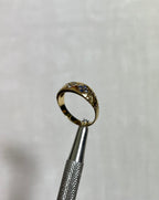 A gold ring with small blue and clear stones, held in the jaws of silver tweezers against a white textured background.