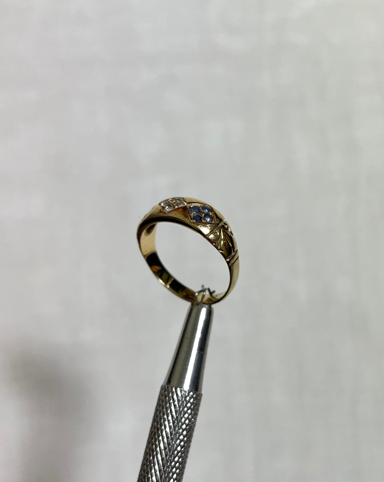 A gold ring with small blue and clear stones, held in the jaws of silver tweezers against a white textured background.